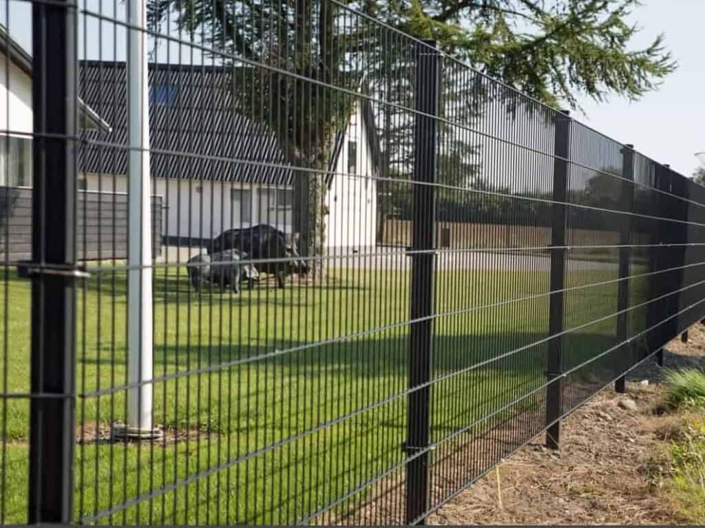 Detalje af hegnstråd på et panelhegn med et hvidt hus og træer i baggrunden fra GA Teknik.